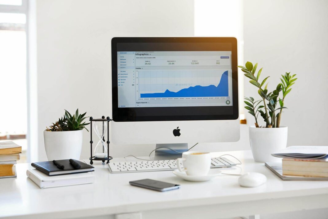 Verslo tendencijos 2025: modernaus darbo stalo vaizdas su iMac kompiuteriu, rodančiu verslo analitikos grafiką, šalia augalai ir kavos puodelis minimalistinėje aplinkoje