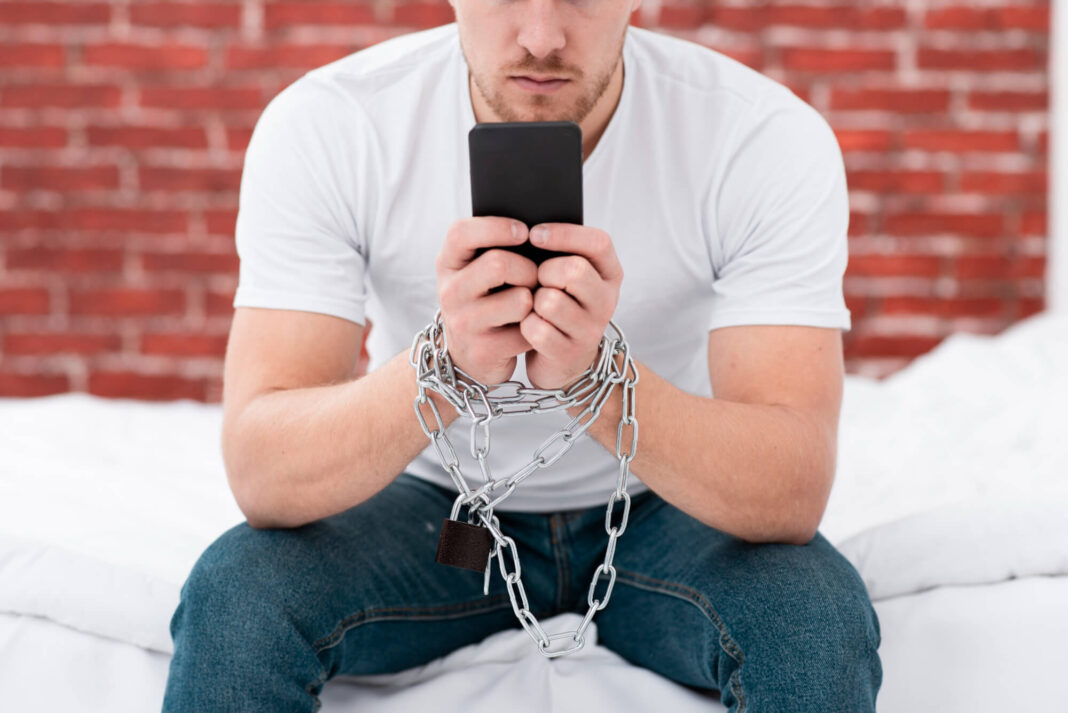 Priklausomybė nuo telefono – vyras su grandinėmis surakintomis rankomis, laiko telefoną.