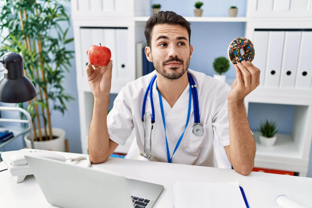Gydytojas, rankose laikantis obuolį ir spurgą, svarsto sveikesnius pasirinkimus, kurie galėtų veikti kaip liaudiški vaistai nuo cholesterolio.