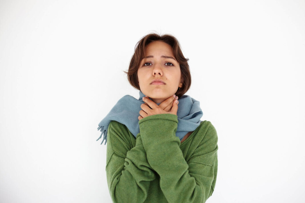 Liaudiški patarimai nuo užkimimo – moteris su šaliku, rankomis laikanti gerklę, rodanti diskomfortą dėl balso praradimo.