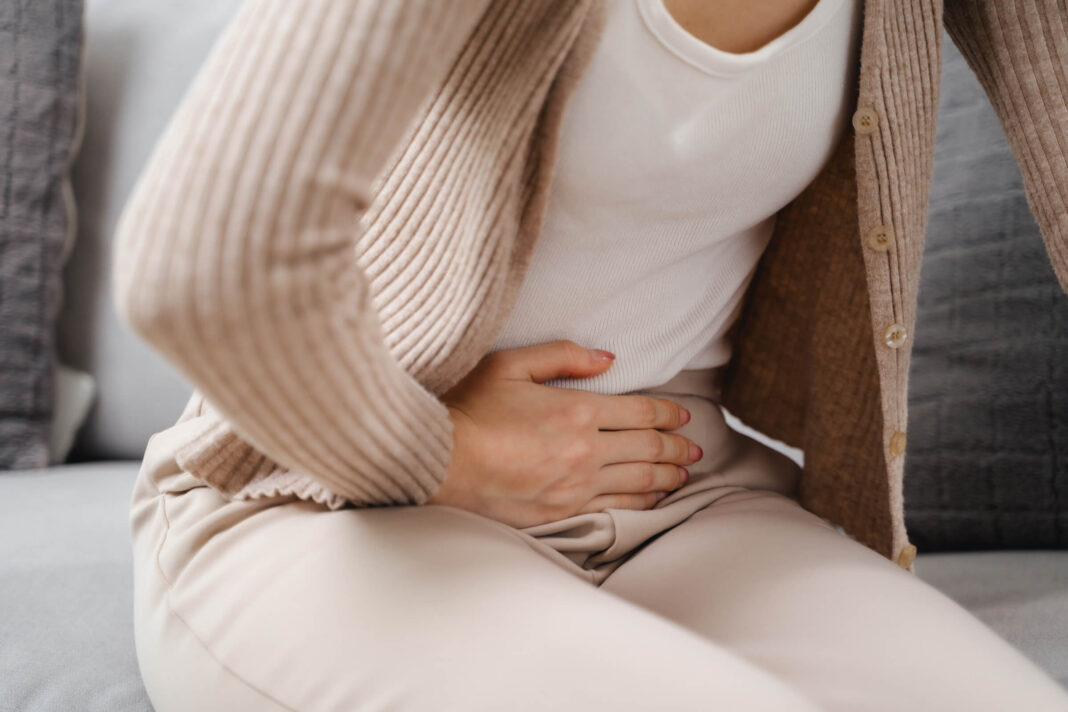 Skrandžio skausmai nerviniu pagrindu, kai moteris sėdi ant sofos ir ranka spaudžia pilvą jausdama diskomfortą.