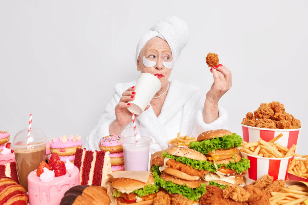 Vyresnio amžiaus moteris su chalatu priešais gausų nesveiko maisto stalą – maistas didinantis kraujospūdį.