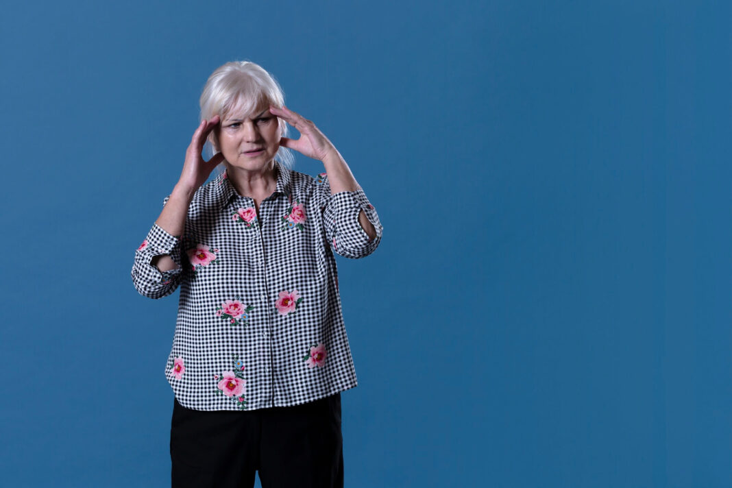 Patarimai nuo galvos svaigimo – vyresnė moteris jaučia diskomfortą ir laikosi už galvos, fonas mėlynas, simbolizuojantis sveikatos temas