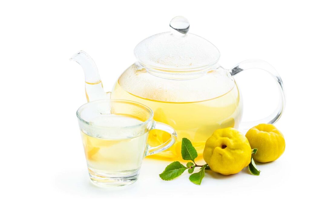 Skaidri stiklinė ir arbatinukas su šilta svarainių arbata baltame fone.