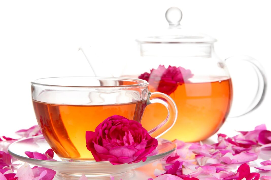 Rožių arbata organizmui – puodelis ir arbatinukas su rožių žiedlapiais, šviesiame fone