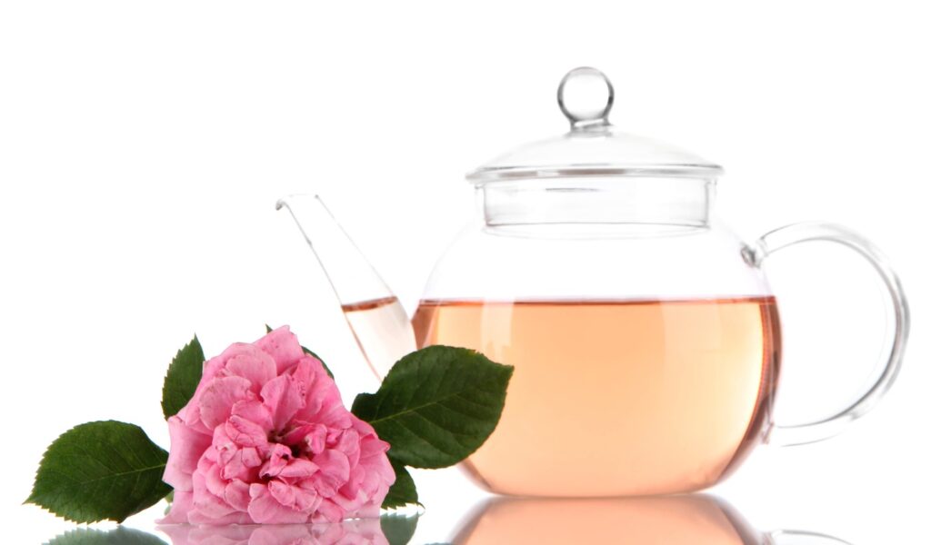 Rožių arbata organizmui – natūralus gėrimas stiklinėje arbatinuke, šalia šviežia rožė su žaliais lapais
