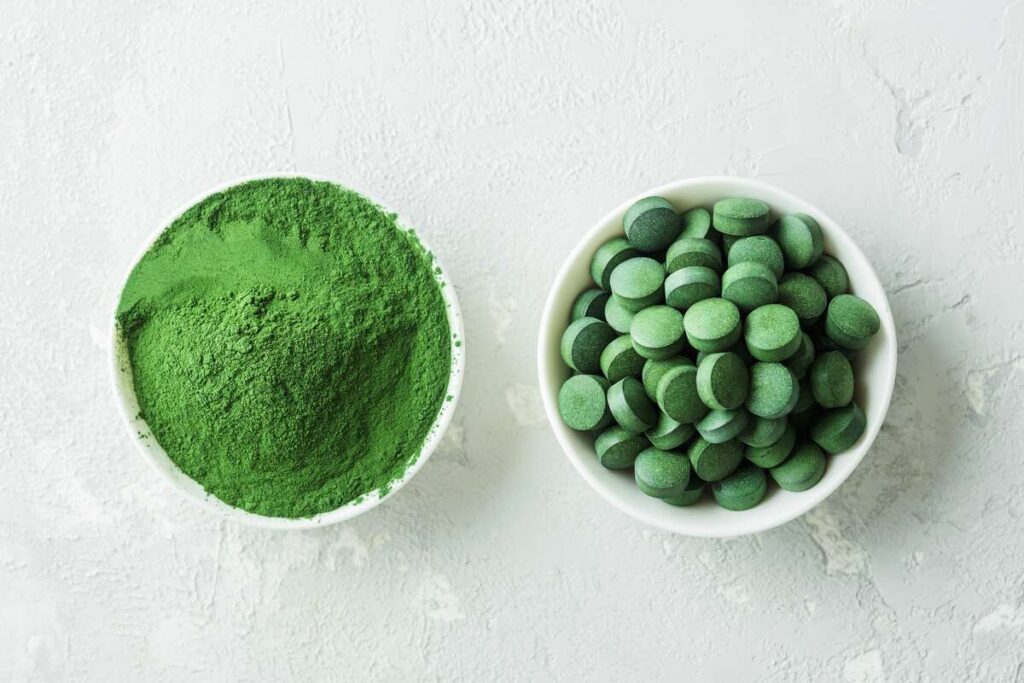 Spirulina milteliai ir tabletės baltuose dubenėliuose ant šviesaus paviršiaus
