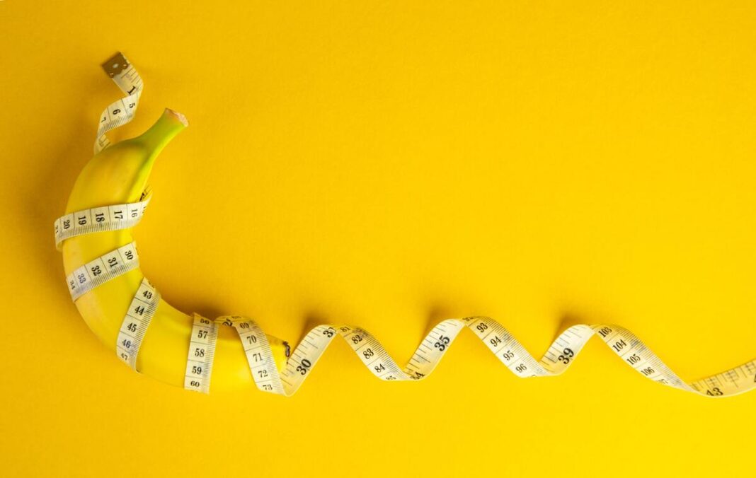 Bananai ir svorio metimas – ant geltono fono pavaizduotas bananas, apjuostas matavimo juosta kaip simbolis dietai ir kūno formoms.