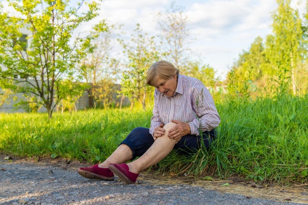 Lapai nuo sąnarių skausmo – moteris jaučia stiprų kelio skausmą, sėdi ant kelio pakraštyje, ieško palengvėjimo.