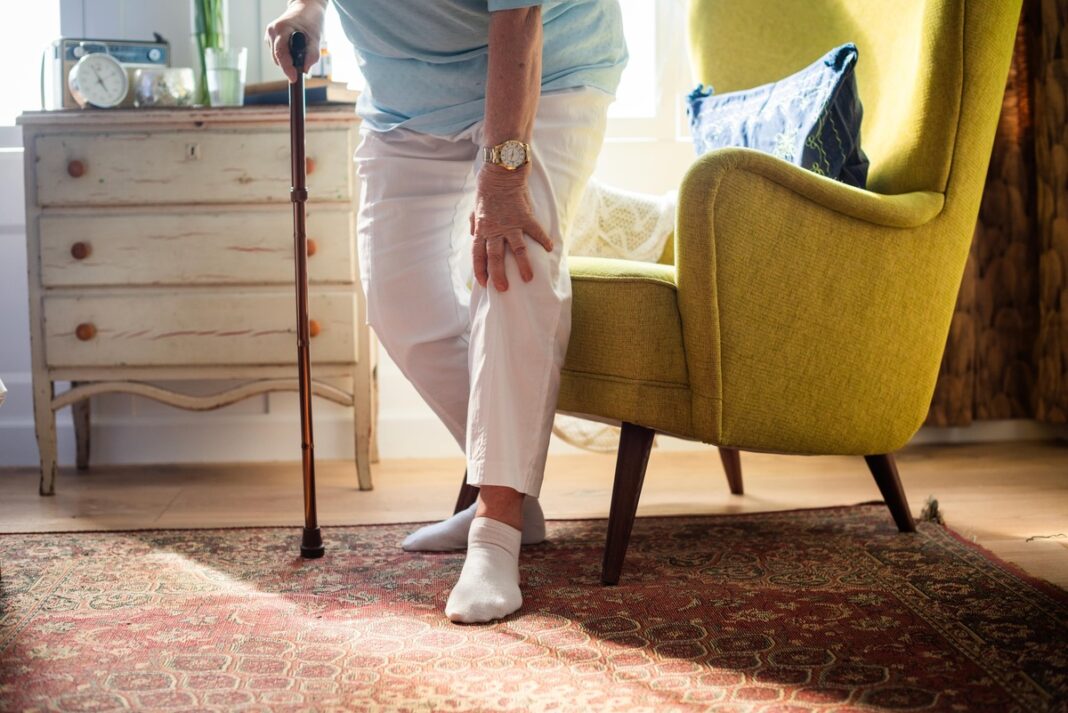 Lauro lapas kojinėse – moteris su lazdele jaučia skausmą kojoje prie krėslo.