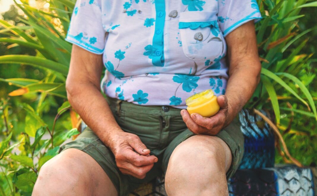 Liaudiškas tepalas iš gvazdikėlių – senjorė lauke tepa tepalą ant skaudančio kelio, naudodama natūralų, naminį preparatą.