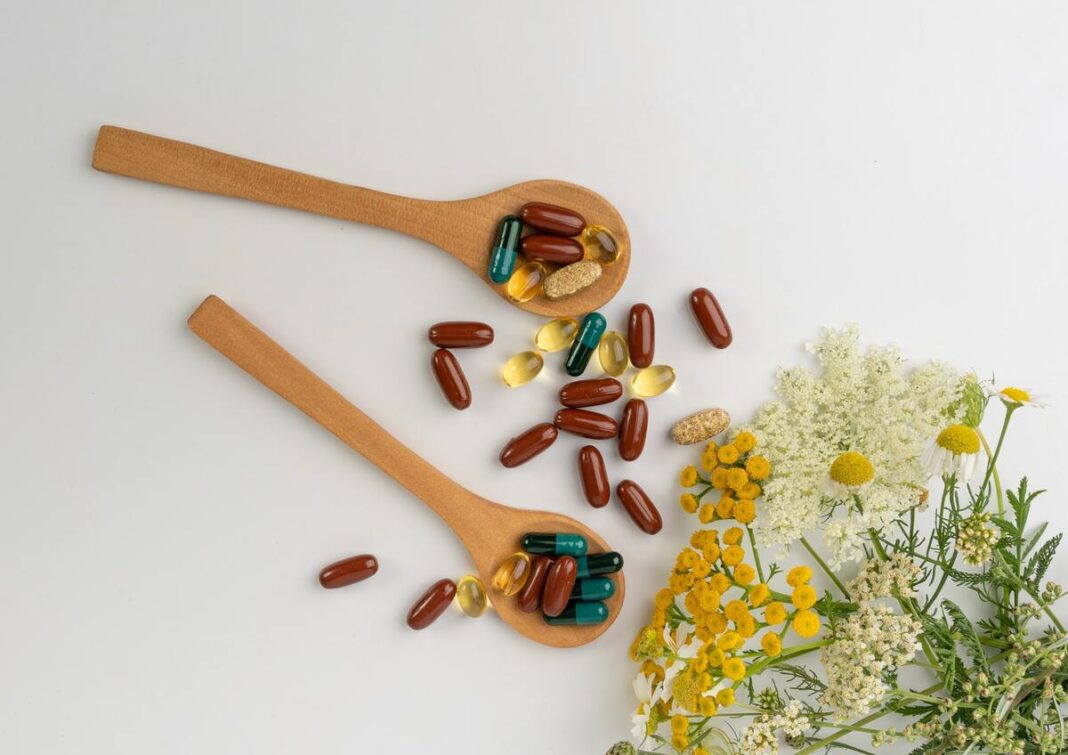 papildų vartojimo laikas – mediniai šaukštai su įvairių rūšių kapsulėmis šalia vaistinių augalų.