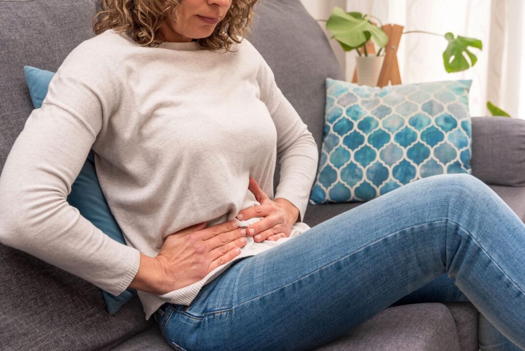 Žarnynas streikuoja – moteris ant sofos laiko rankas ant pilvo dėl diskomforto ir virškinimo sutrikimų.