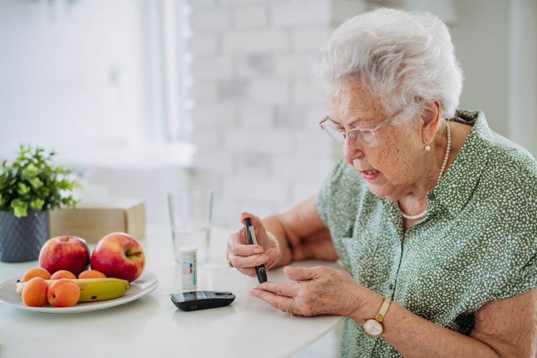 Senjorė matuojasi gliukozės kiekį kraujyje, šalia ant stalo guli vaisiai – iliustracija apie bulvyčių fri ir diabeto ryšį.