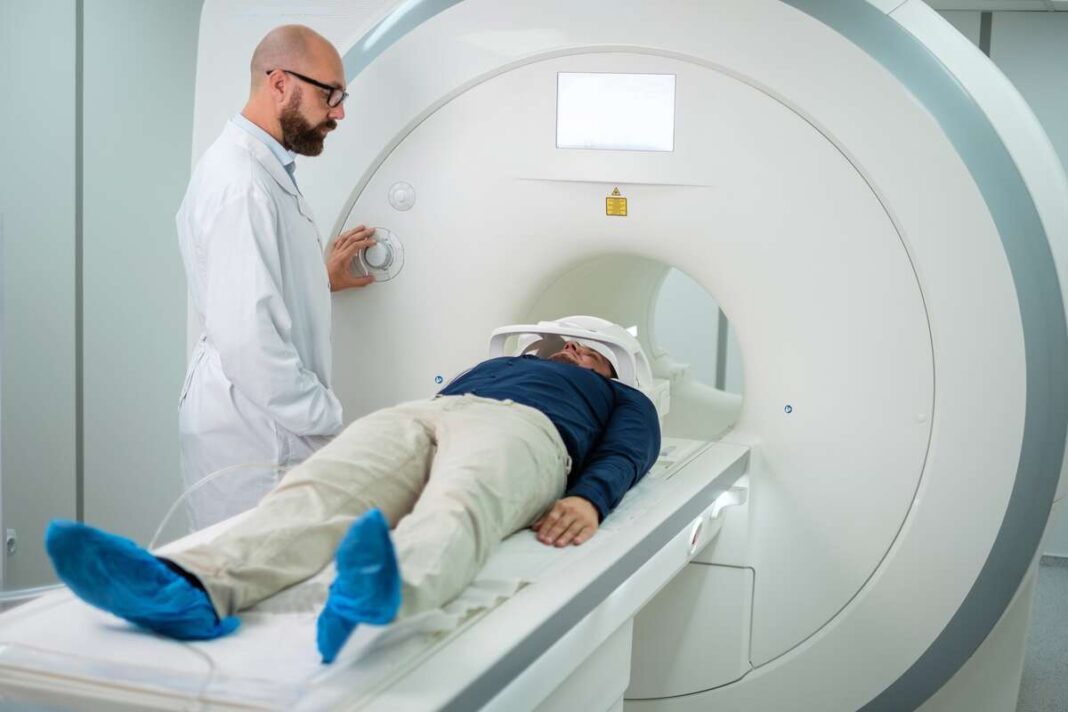 Pacientas guli MRT aparate, o šalia stovi gydytojas, stebintis tyrimo eigą.