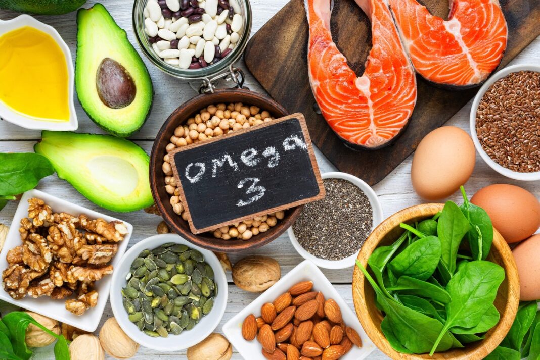 Įvairūs geriausi omega-3 šaltiniai – avokadas, lašiša, riešutai, sėklos, špinatai ir kiti sveiki produktai ant medinio stalo.