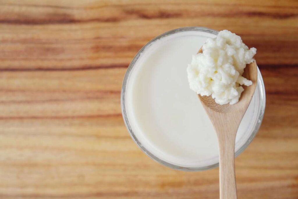 Kefyras ar jogurtas – stiklinė kefyro su mediniu šaukštu ir kefyro grūdeliais ant medinio stalo.