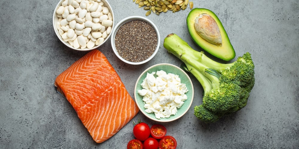 Lašiša, avokadas, brokoliai, chia sėklos, pupelės ir kiti geriausi omega-3 šaltiniai ant pilko stalo.