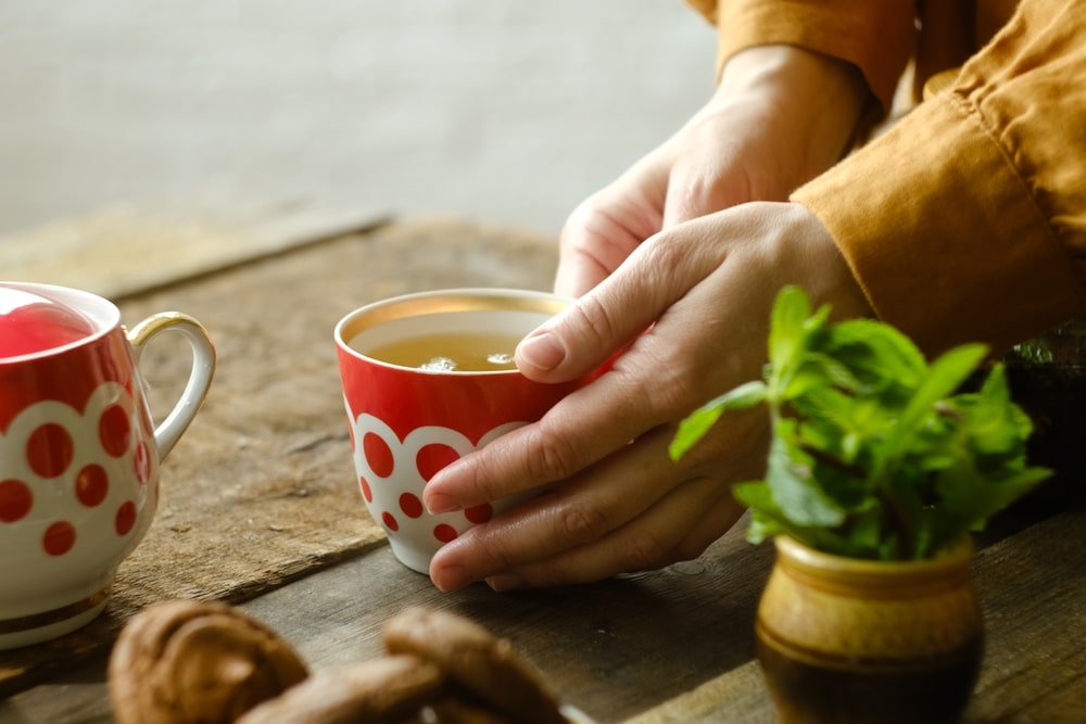 Raudonuose puodeliuose patiekiama karšta pavojinga arbata – rankose laikomas gėrimas, kuris gali kelti netikėtą riziką sveikatai.