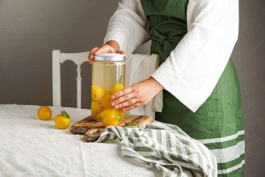 Geltonos slyvos užpiltos kompotu stiklainyje, ruošiamos namuose ant stalo su medine lentele.