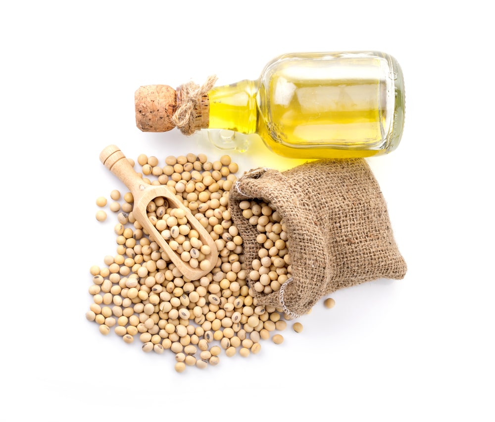 Stiklinis butelis su sojų aliejumi ir šalia paberta sauja sojų pupelių ant lino maišelio – vienas iš daugiausia omega-6 turinčių aliejų šaltinių.