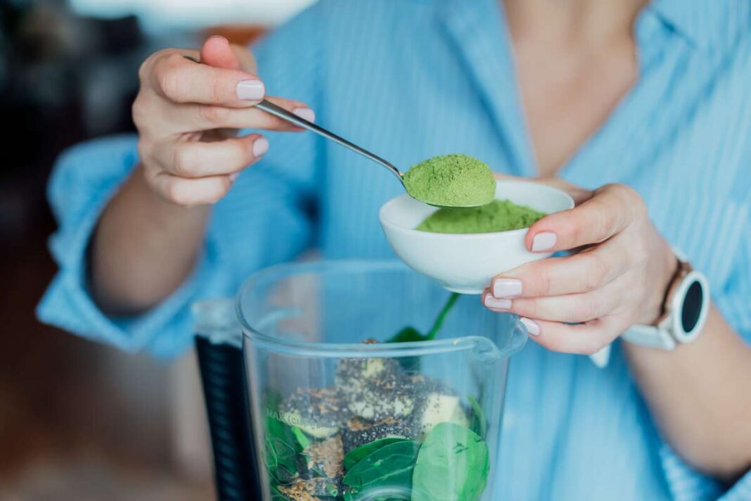 spirulinos-nauda-sveikatai-milteliai-smoothie Moteris į kokteilį deda šaukštą spirulinos miltelių – natūralus būdas stiprinti sveikatą ir valyti organizmą.