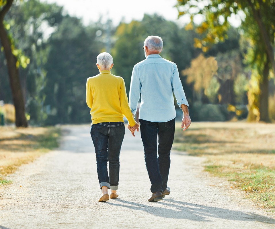 Vaikščiojimas kartu – pagyvenusi pora eina susikibusi rankomis saulėtame take.