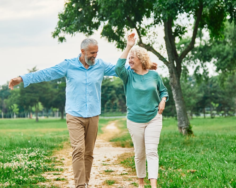 Vyresnio amžiaus pora šypsodamasi vaikšto parke – vaikščiojimas nuo depresijos padeda išlaikyti gerą nuotaiką ir emocinę pusiausvyrą.