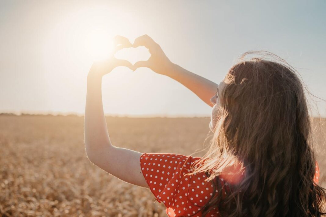 vitamino D šaltiniai be tablečių – moteris laukuose rankomis formuoja širdį, pro ją sklinda saulės spinduliai.