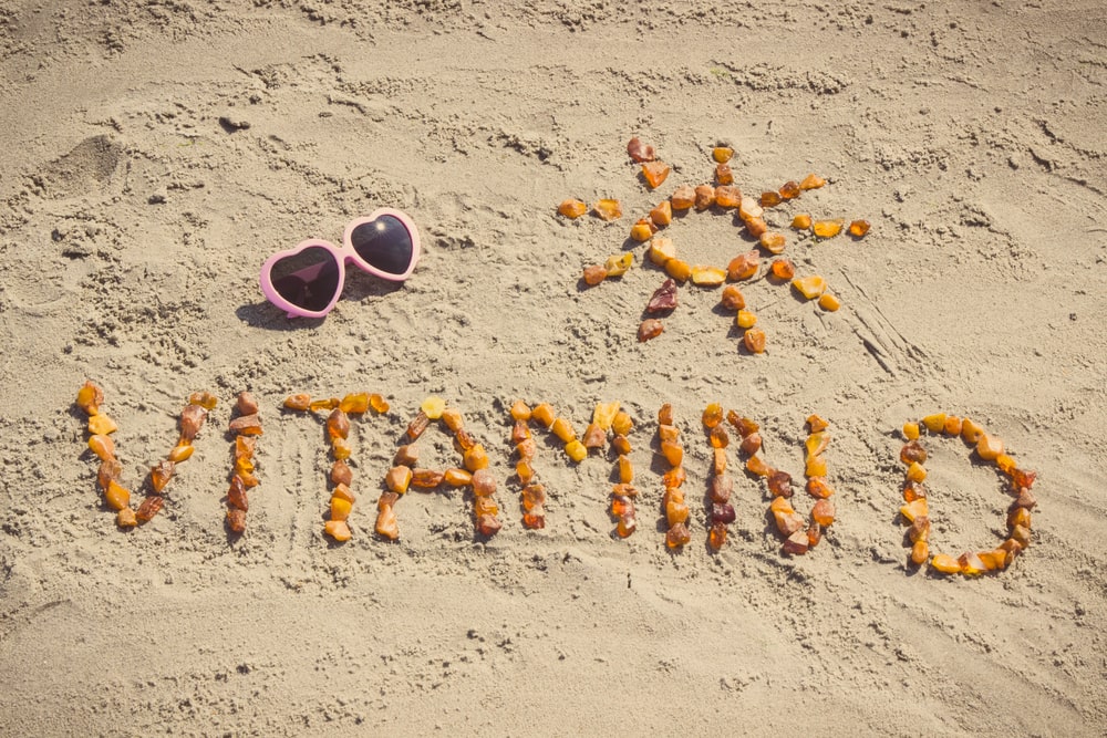 vitamino D šaltiniai be tablečių – smėlyje gintaru sudėliotas užrašas su saulės simboliu ir rožiniais širdies formos akiniais nuo saulės.