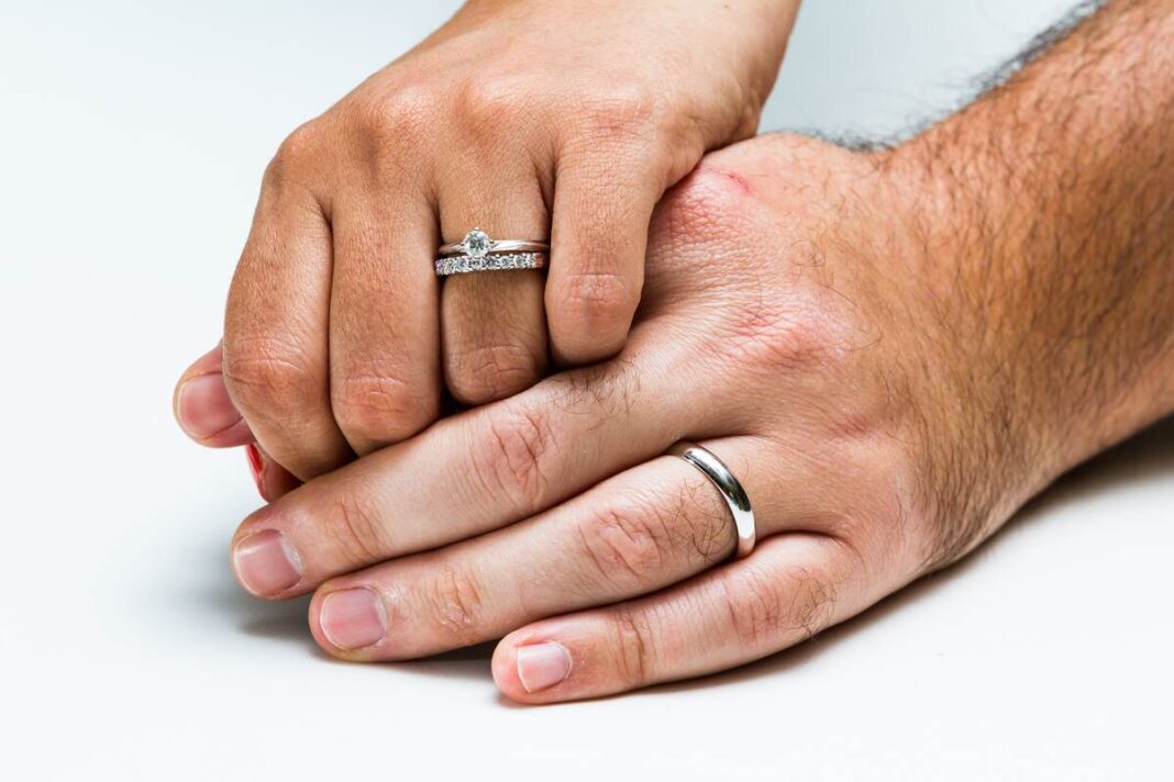 žiedai su deimantais – moters ranka su deimantine juostele ir sužadėtuvių žiedu šalia vyro vestuvinio žiedo.