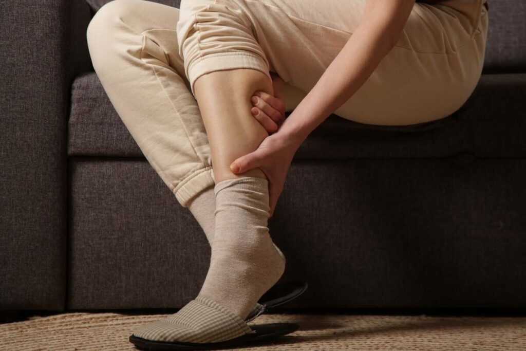 Blauzdos raumenys ir kraujotaka – moteris jaučia įtampą kojose ir masažuoja blauzdą sėdėdama ant sofos.