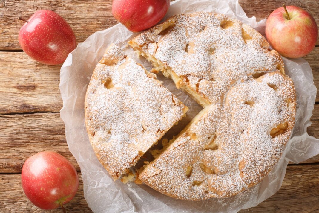 Apvalus senovinis obuolių pyragas, padalintas gabalėliais ir apibarstytas cukraus pudra, šalia keli raudoni obuoliai.