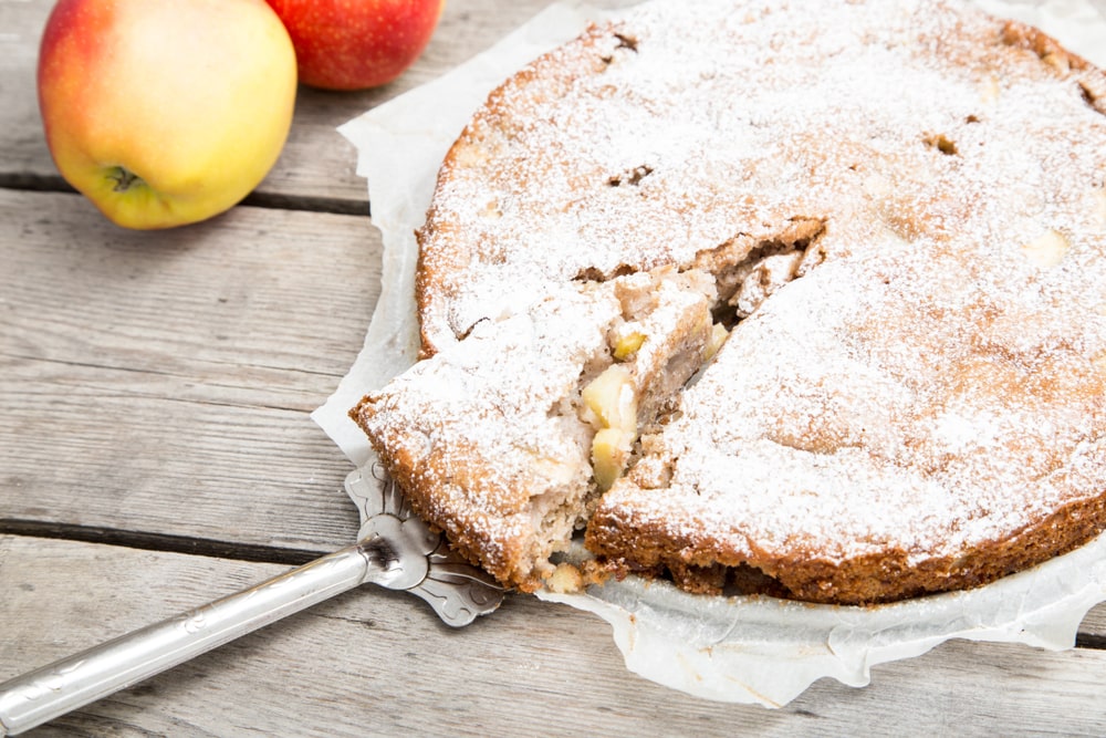 Senovinis obuolių pyragas, apibarstytas cukraus pudra, padėtas ant medinio stalo su šviežiais obuoliais šalia.