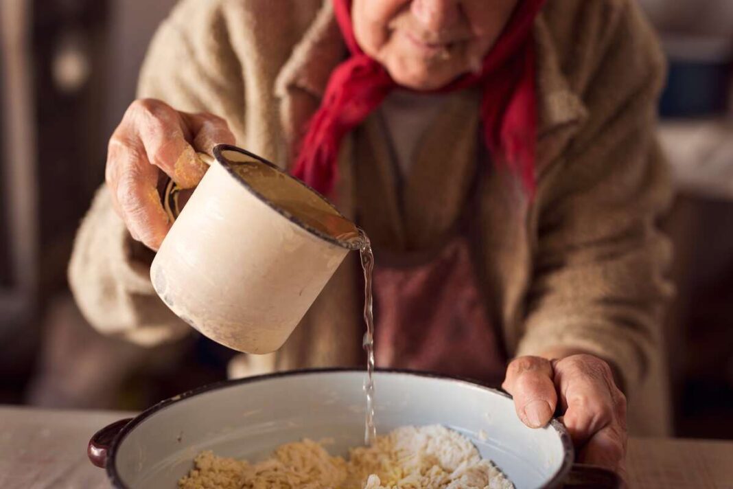 Močiutės rankos ruošia paprastą sveikatos receptą – vanduo pilamas ant natūralių ingredientų dubenyje.