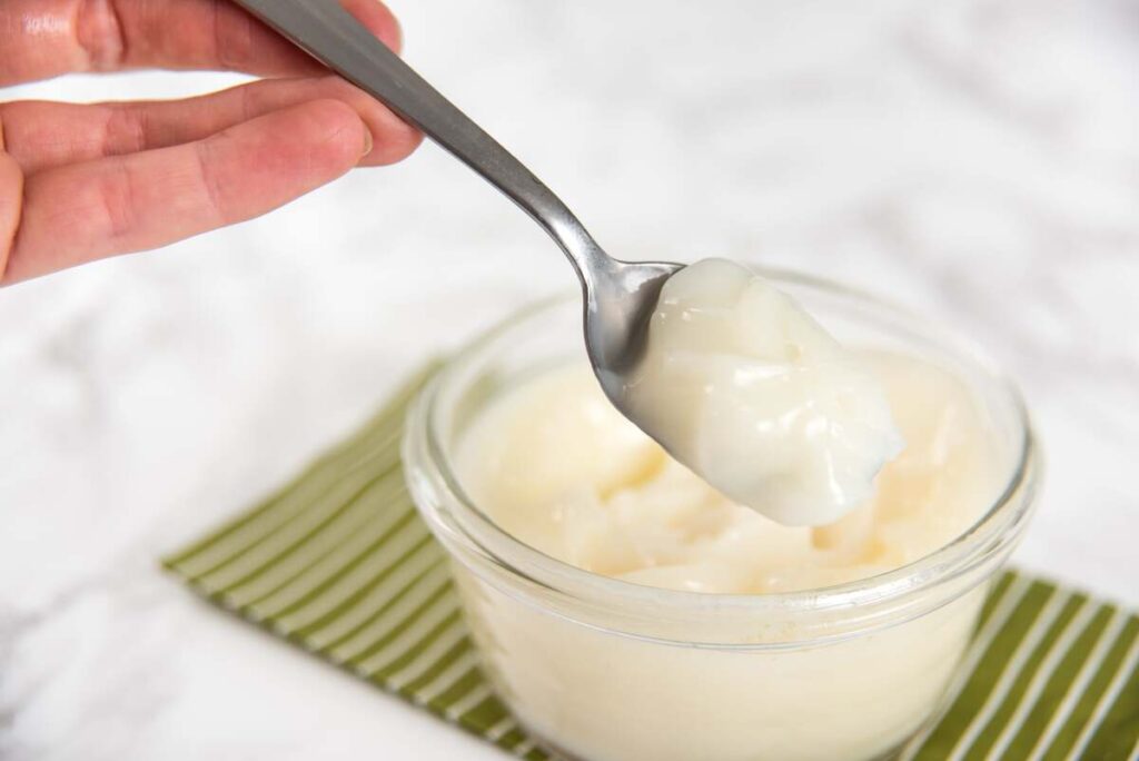 žąsų taukai šaukšte virš stiklinio indo – natūralūs, švelnūs ir lengvai naudojami virtuvėje bei sveikatai.