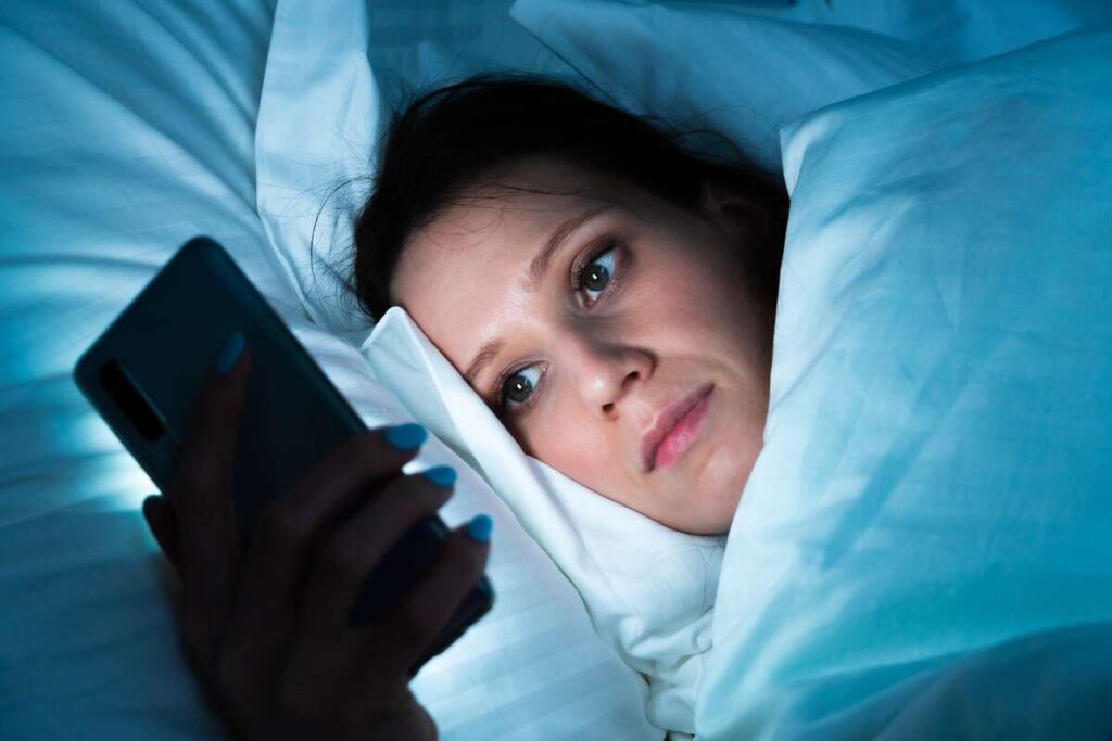 Ar melatoninas kenkia sveikatai – moteris naktį žiūri į telefoną, patirianti miego sutrikimus dėl šviesos poveikio.