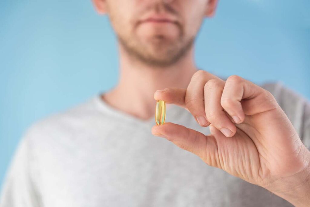 Kada verta gerti vitaminą D – vyras rankoje laiko kapsulę su vitaminu D prieš mėlyną foną.