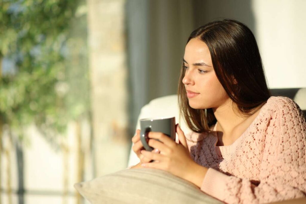 Kava tuščiu skrandžiu – moteris ryte žiūri pro langą, rankose laikydama kavos puodelį.