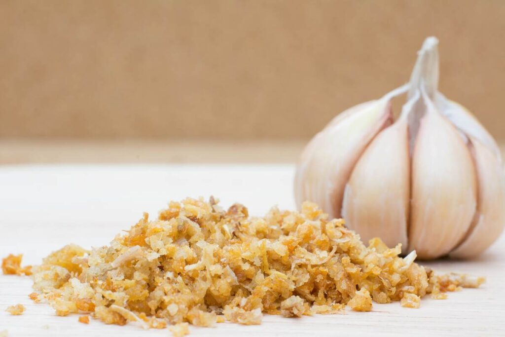 Keptas česnakas naudingas sveikatai – susmulkintas, traškus česnakas šalia šviežios galvutės.