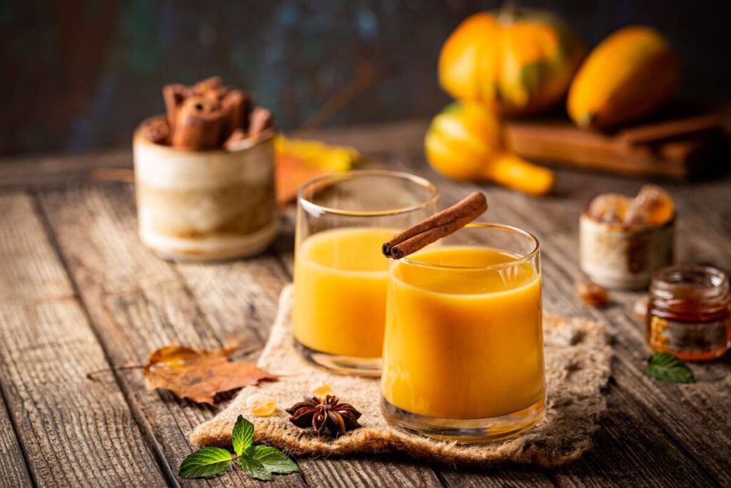 Moliūgų kokteilis cholesteroliui mažinti stiklinėje su cinamono lazdele ant medinio stalo.