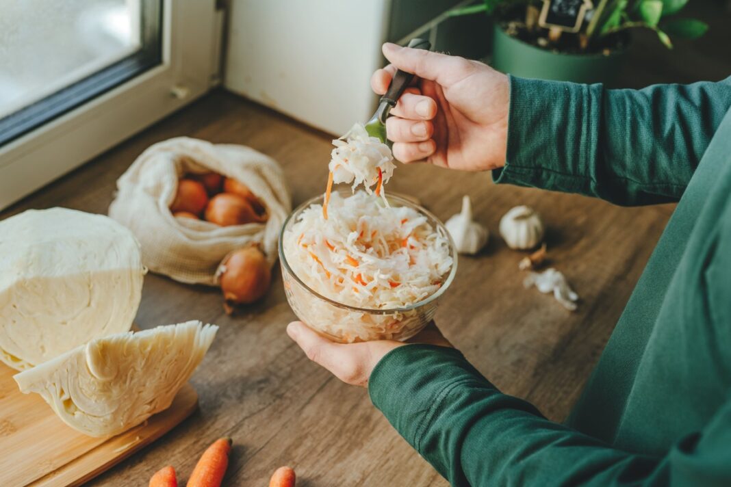 raugintu-kopustu-poveikis-naturali-fermentacija raugintų kopūstų poveikis – žmogus laiko dubenį šviežiai raugintų kopūstų su morkomis virtuvėje.