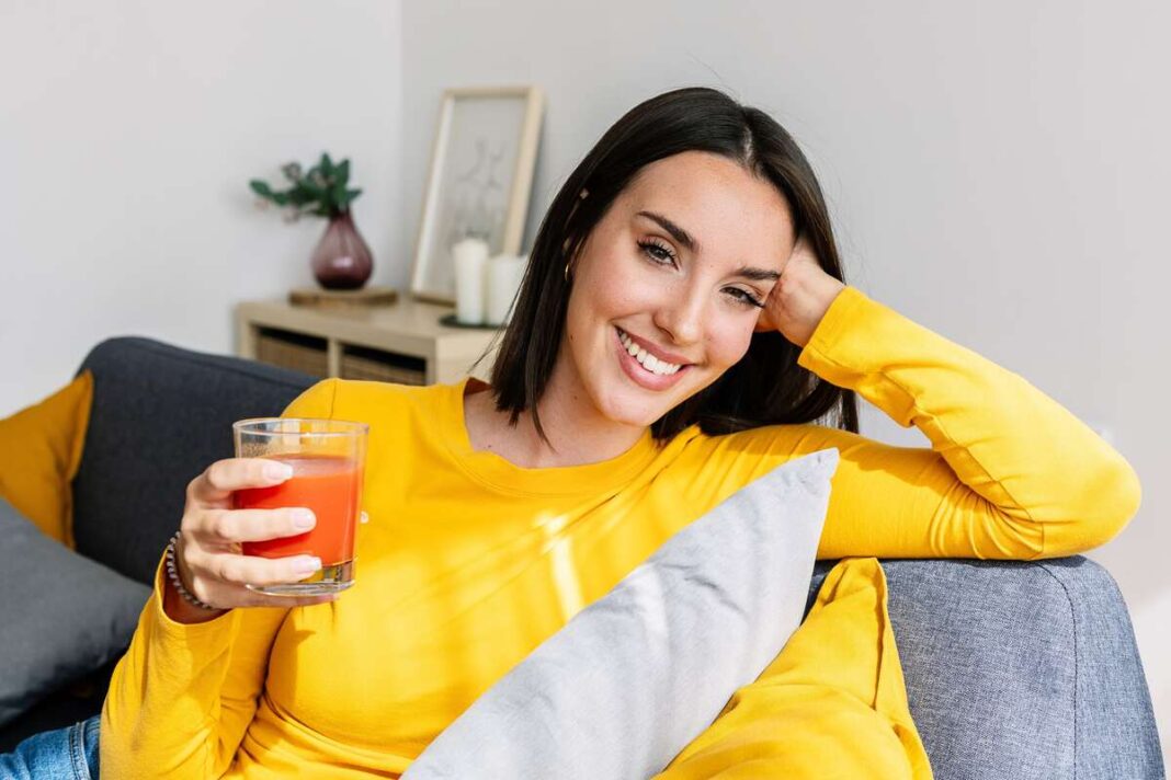 Sultys širdžiai ir kraujagyslėms – moteris laisvai sėdi ant sofos ir šypsodamasi geria pomidorų sultis.