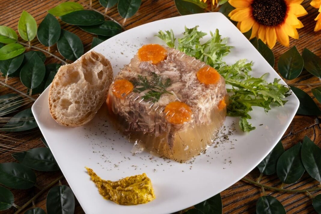 Tradicinė šaltiena su morkomis, žalumynais ir duonos riekėle, patiekta ant balto porcelianinio indo.