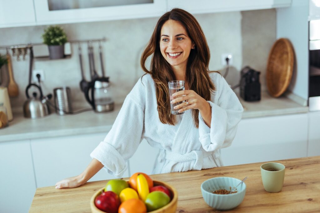 Arbata kepenims – moteris ryte virtuvėje geria stiklinę vandens, pradėdama dieną sveikatingai.