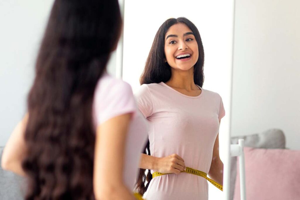 kaip numesti svorio sveikai – jauna moteris matuojasi liemenį ir šypsosi žiūrėdama į veidrodį