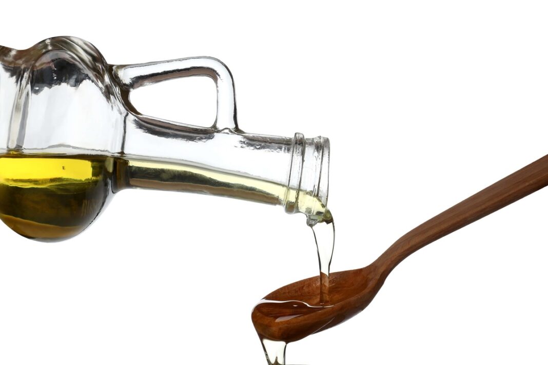 linų sėmenų aliejus po ligos pilamas iš stiklinio butelio į medinį šaukštą baltame fone.