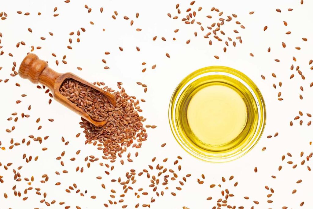 linų sėmenų aliejus po ligos – stiklinis indas su aliejumi ir šaukštas pilnas linų sėklų ant šviesaus fono.
