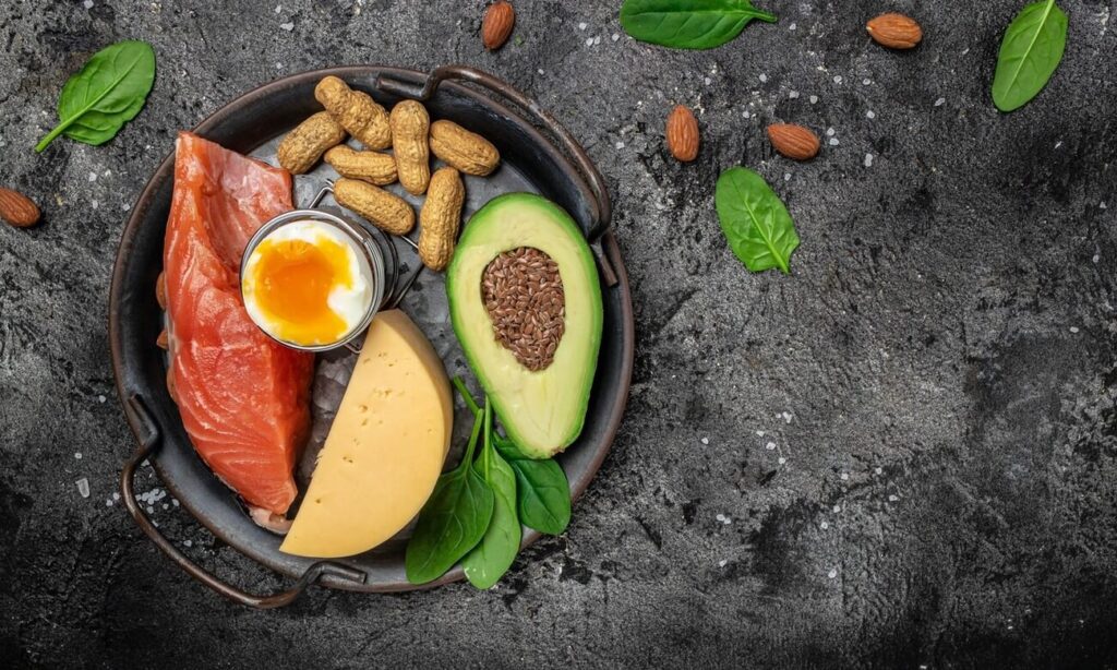 Mažinti cholesterolį be vaistų padeda sveikų riebalų produktai: avokadas, lašiša, kiaušinis, riešutai ir sėklos vienoje lėkštėje.