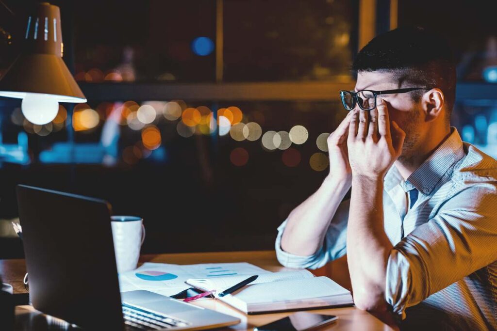 naktinio darbo pasekmės sveikatai – vyras prie kompiuterio naktį jaučiasi pavargęs, kenčia nuo akių įtampos ir miego trūkumo.